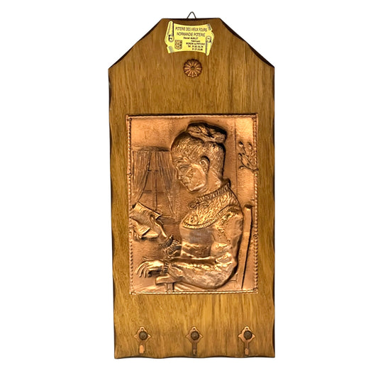 A wooden wall hanging plaque featuring a raised relief carving of a seated woman in traditional dress, with three metal hooks at the bottom and a small gold-colored hanging tag at the top.