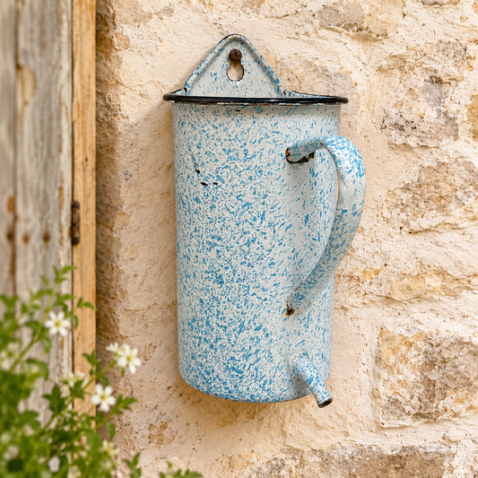 A vintage-style light blue and white speckled enamel wall-mounted watering can with a black rim, shown mounted on a rustic beige stone wall.