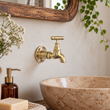 A rustic bathroom scene featuring a round beige stone vessel sink, a vintage brass wall-mounted faucet, a wooden countertop, and decorative toiletries including a soap dispenser, soap bar, and dried flowers.
