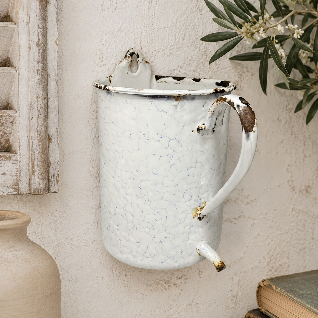 French vintage enamel planter jug or kitchen pot with a handle and spout, perfect for a rustic garden, displayed on a wall with a plant and a shutter in the background.