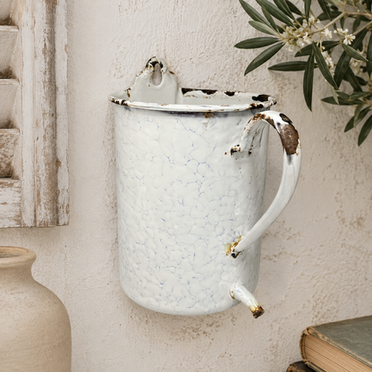 French vintage enamel planter jug or kitchen pot with a handle and spout, perfect for a rustic garden, displayed on a wall with a plant and a shutter in the background.
