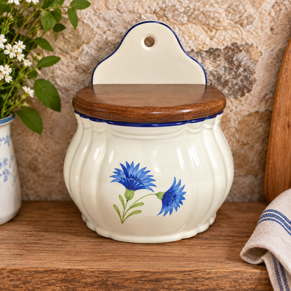 Vintage French wall-mounted ceramic salt box with removable wooden lid, displayed on a rustic wooden kitchen worktop against a warm natural stone wall. Cream glazed fluted ceramic body featuring hand-painted blue cornflowers, green stems and dark blue trim, with a curved hanging backplate. Styled with white flowers in a blue-and-white ceramic jug, a wooden board and blue-striped linen. Authentic French brocante kitchenware, made in France, ideal for rustic farmhouse and French country kitchens.