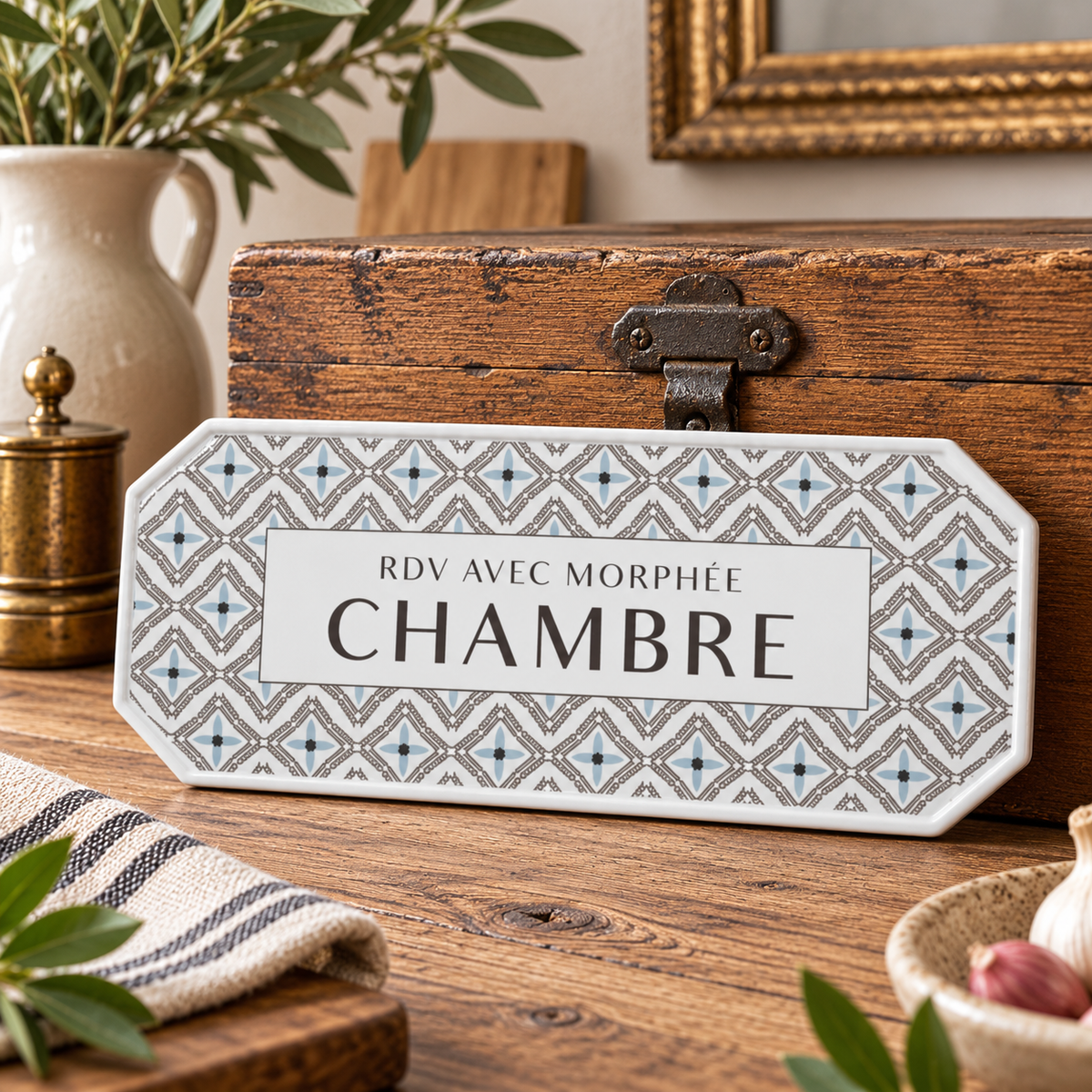 A decorative French bedroom room sign with a geometric patterned border and a white center panel reading 'RDV AVEC MORPHÉE CHAMBRE', displayed on a wooden tabletop with rustic home decor props.