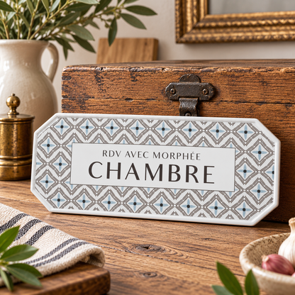 A decorative French bedroom room sign with a geometric patterned border and a white center panel reading 'RDV AVEC MORPHÉE CHAMBRE', displayed on a wooden tabletop with rustic home decor props.