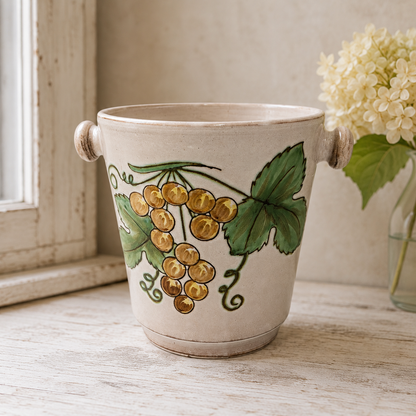 French hand-painted glazed champagne bucket adorned with grape motifs, placed on a wooden table beside a flower in a vase near a window, perfect for a wedding setting or a stylish home bar.