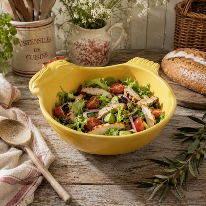 A yellow ceramic salad bowl in a chicken shape, filled with a fresh salad, set on a rustic wooden table with a wooden spoon, a striped cloth, and bread in the background.