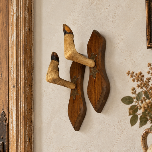 Pair of authentic vintage French deer hoof taxidermy coat hooks mounted on individual shaped wooden plaques, displayed on a textured cream plaster wall in a rustic French country house hallway. Each hook features natural golden-brown deer fur, dark hooves and decorative metal mounting plates, with aged wooden plaques showing their original patina and character.  