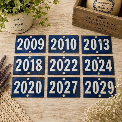 Collection of vintage French blue vitreous enamel number plaques displayed on a rustic wooden tabletop with lavender, a weathered wooden crate and trailing greenery. Authentic 1960s railway-style number signs arranged in a French country brocante setting.