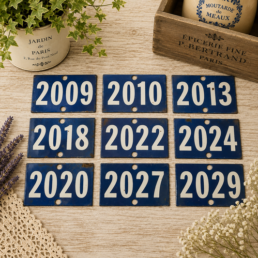 Collection of vintage French blue vitreous enamel number plaques displayed on a rustic wooden tabletop with lavender, a weathered wooden crate and trailing greenery. Authentic 1960s railway-style number signs arranged in a French country brocante setting.
