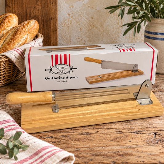 A wooden and metal bread scimitar set displayed on a rustic wooden table, with its white and red 'Bistron Cafe' retail box placed behind it. The set includes a wooden handle, a stainless steel blade, and guide rods, with decorative bread, a striped cloth, and a potted plant in the background.