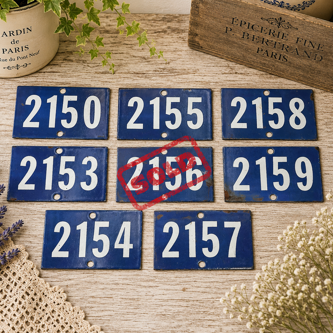 Collection of vintage French blue vitreous enamel number plaques displayed on a rustic wooden tabletop with lavender, a weathered wooden crate and trailing greenery. Authentic 1960s railway-style number signs arranged in a French country brocante setting.