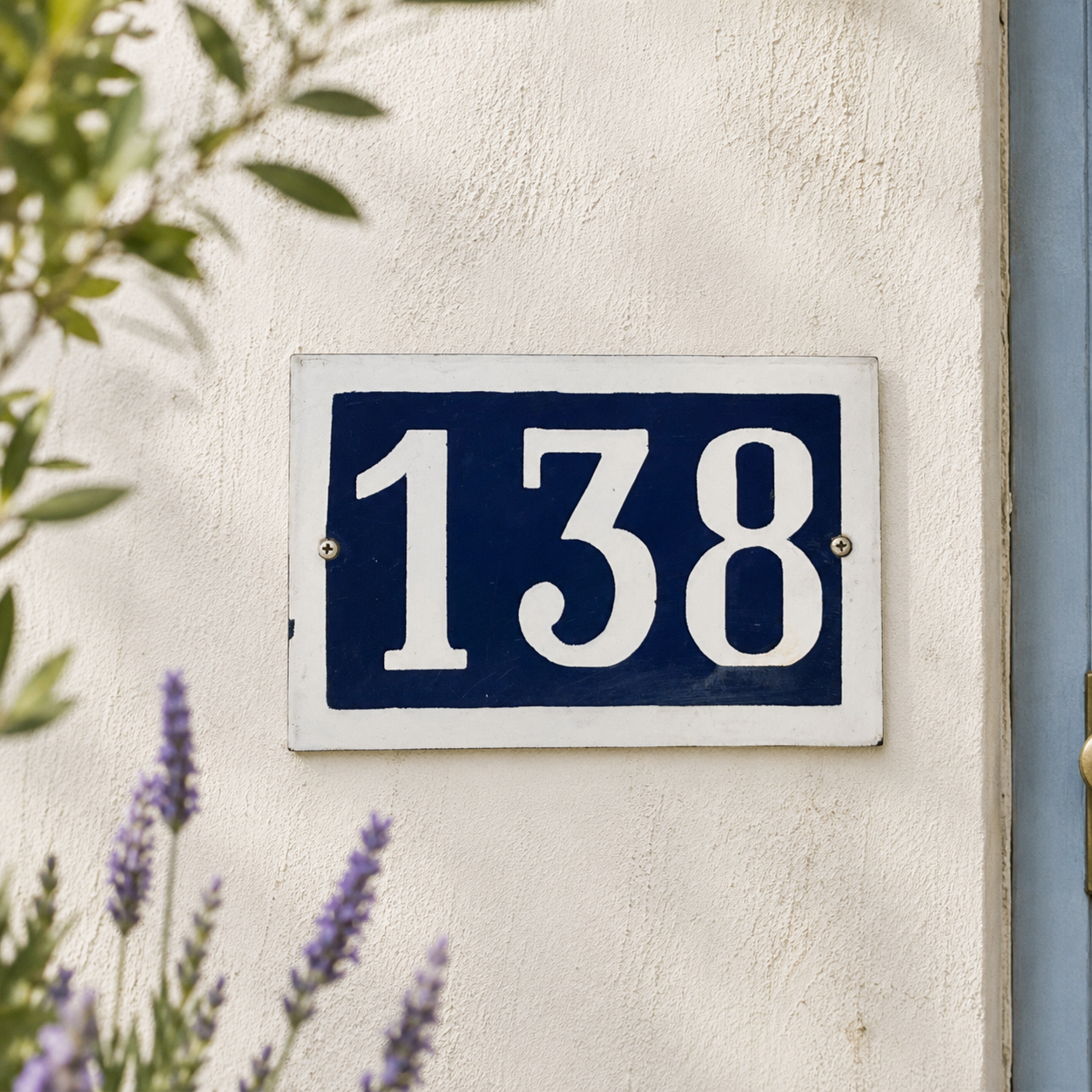 House number '138' on a blue and white sign mounted on a beige wall with plants in the background.
