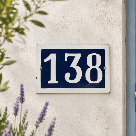 House number '138' on a blue and white sign mounted on a beige wall with plants in the background.