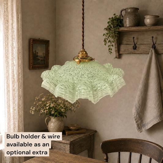 A light green ruffled glass pendant light hangs over a wooden dining table in a rustic room with a textured beige wall, a wooden shelf, and a hanging towel. A text overlay in the bottom left corner reads 'Bulb holder & wire available as an optional extra'.