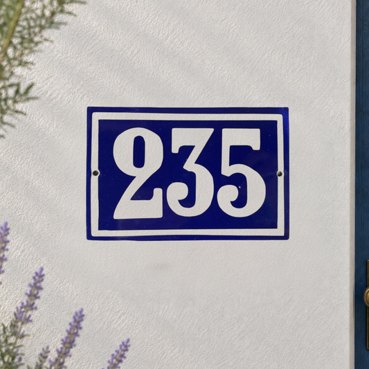 A blue and white enamel house number plaque reading '235' mounted on a textured white stucco wall, with soft green and purple plants framing the left side.