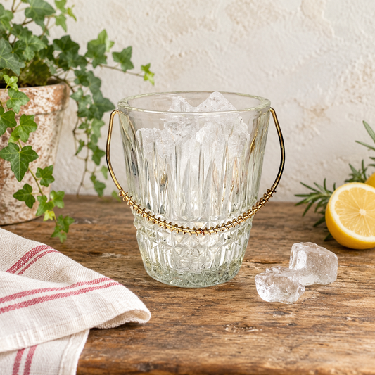 French small ice bucket on a wooden counter top with some ice cubes and a lemon beside a tea cloth and a plant
