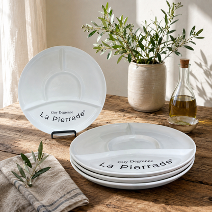 Three white ceramic La Pierrade Guy Degrenne divided plates arranged on a rustic wooden table with olive branches, a vase, and a glass bottle in a styled setting.