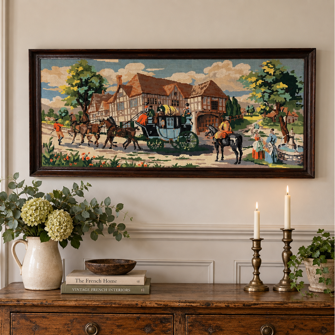 A framed horizontal needlepoint depicting a classic village scene with horse-drawn carriages and Tudor-style houses, displayed on a cream wall above a rustic wooden console table decorated with a vase of greenery and lit candles.