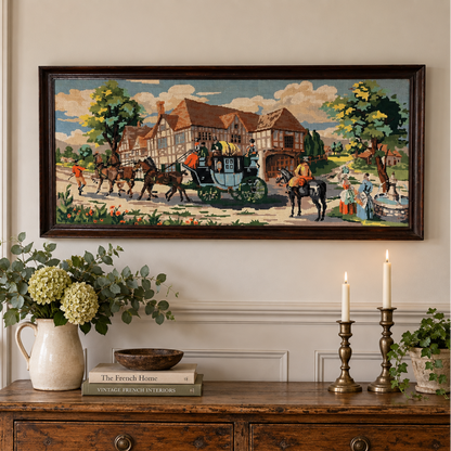 A framed horizontal needlepoint depicting a classic village scene with horse-drawn carriages and Tudor-style houses, displayed on a cream wall above a rustic wooden console table decorated with a vase of greenery and lit candles.