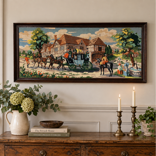 A framed horizontal needlepoint depicting a classic village scene with horse-drawn carriages and Tudor-style houses, displayed on a cream wall above a rustic wooden console table decorated with a vase of greenery and lit candles.