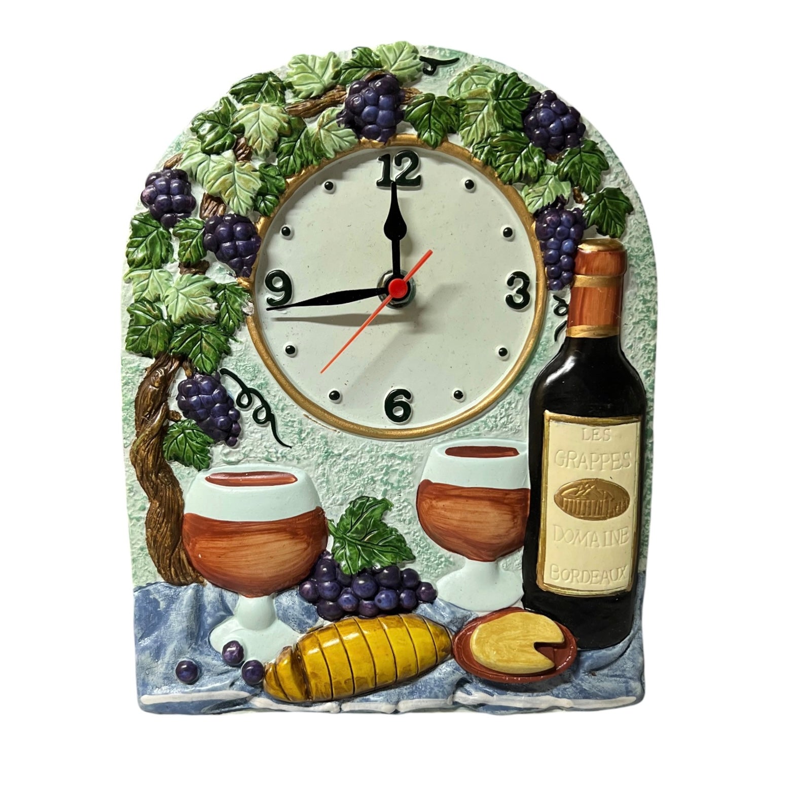 A decorative wine-themed wall clock with a sculpted face featuring grapes, a wine bottle, two wine glasses, cheese, and a blue cloth. The clock hands are black with a red second hand, and the center dial is white with black numbers.