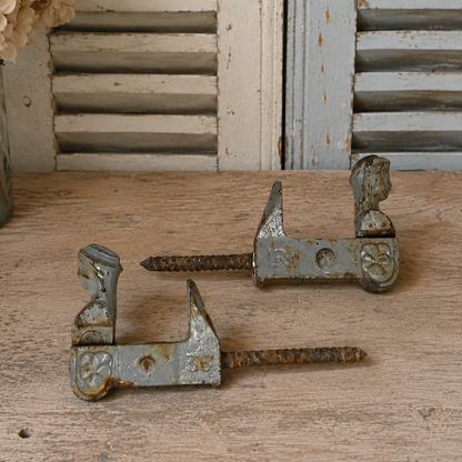 Two vintage rusted metal shutter brackets with long screw elements, displayed on a rustic wooden surface with distressed shutters and dried flowers in the background.