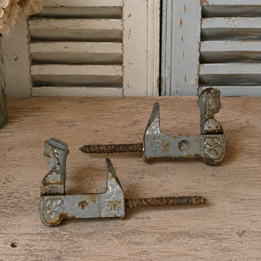 Two vintage rusted metal shutter brackets with long screw elements, displayed on a rustic wooden surface with distressed shutters and dried flowers in the background.
