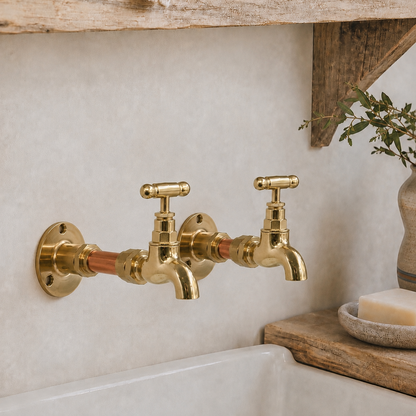 A pair of vintage-style brass wall-mounted kitchen faucets with cross handles and copper accents, installed on a textured beige wall above a white sink, with rustic wooden decor and a soap bar nearby.