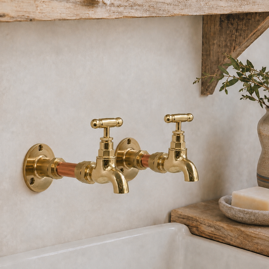A pair of vintage-style brass wall-mounted kitchen faucets with cross handles and copper accents, installed on a textured beige wall above a white sink, with rustic wooden decor and a soap bar nearby.