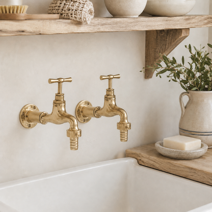 Gold faucet taps on a white sink with a wooden shelf and plant in the background
