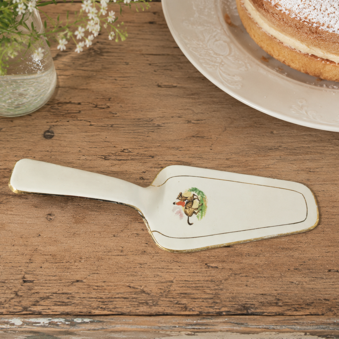 Vintage French porcelain cake server displayed on a rustic wooden table beside a sponge cake and delicate white flowers. The decorative serving piece features a traditional printed motif and gold trim, styled in a warm French country kitchen setting.