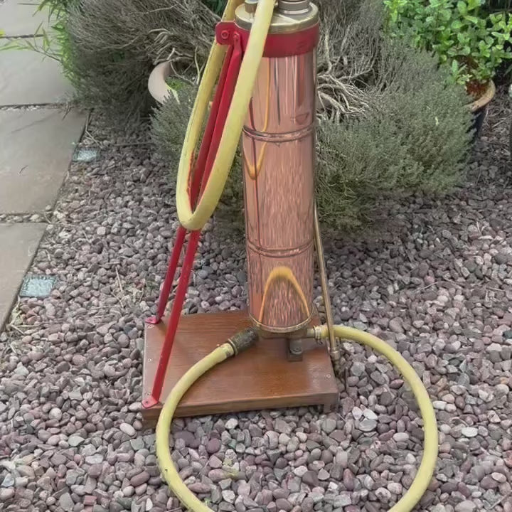 Video of French vintage copper garden sprayer with a yellow hose and nozzle, standing upright on a wooden mount with a red-painted metal bracket. A wooden handle for pumping sits at the top, making it ideal for a vintage garden, all set against a white background.