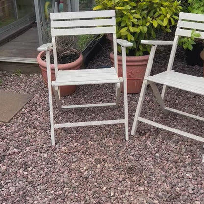 A video featuring a pair of French vintage wooden slatted garden folding chairs set in a charming garden with a greenhouse, potted plants, and a gravel path, perfect for a rustic country cottage vibe.