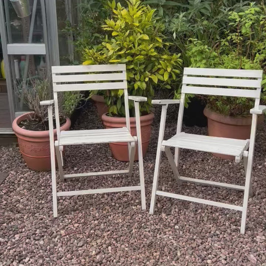 Video of a pair of French vintage wooden slatted garden folding chairs sit in a charming garden setting on a gravel path, perfect for a cosy country cottage garden.