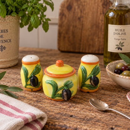 Three hand-painted ceramic salt and pepper shakers and a matching sugar bowl with an olive branch design, styled on a rustic wooden table with a bottle of olive oil, a potted herb, and a striped cloth.