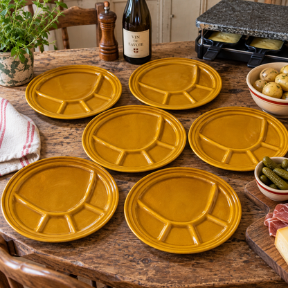 Six mustard yellow ceramic compartment plates arranged on a rustic wooden dining table, surrounded by food props like a bottle of wine, a pepper mill, olives, pickles, and a plant.