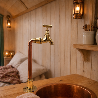 A rustic bathroom or vanity setup featuring a tall copper and brass faucet rising above a matching copper sink, set against a warm wood-paneled wall with lit wall sconces.