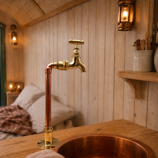 A rustic bathroom or vanity setup featuring a tall copper and brass faucet rising above a matching copper sink, set against a warm wood-paneled wall with lit wall sconces.