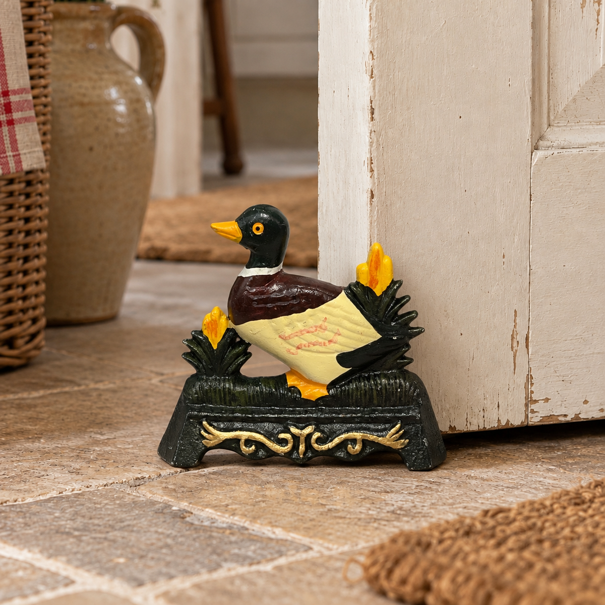 A decorative duck door stop figurine standing on a dark base with yellow flowers, placed on a stone floor beside a white door in a rustic indoor setting.