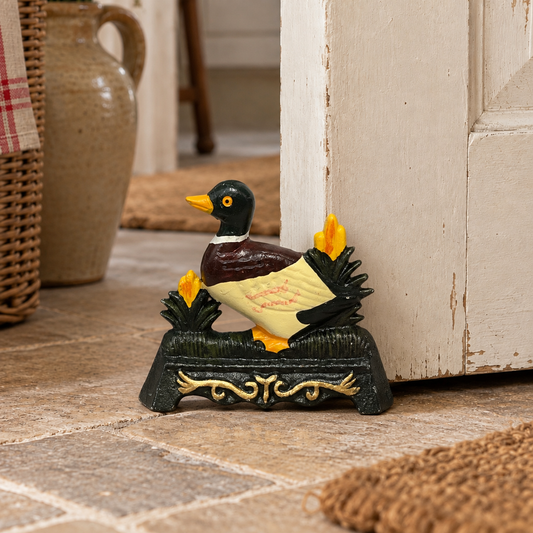 A decorative duck door stop figurine standing on a dark base with yellow flowers, placed on a stone floor beside a white door in a rustic indoor setting.