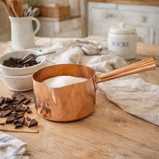 Copper chocolate pan with sugar on a wooden table with chocolate and kitchen utensils in the background.