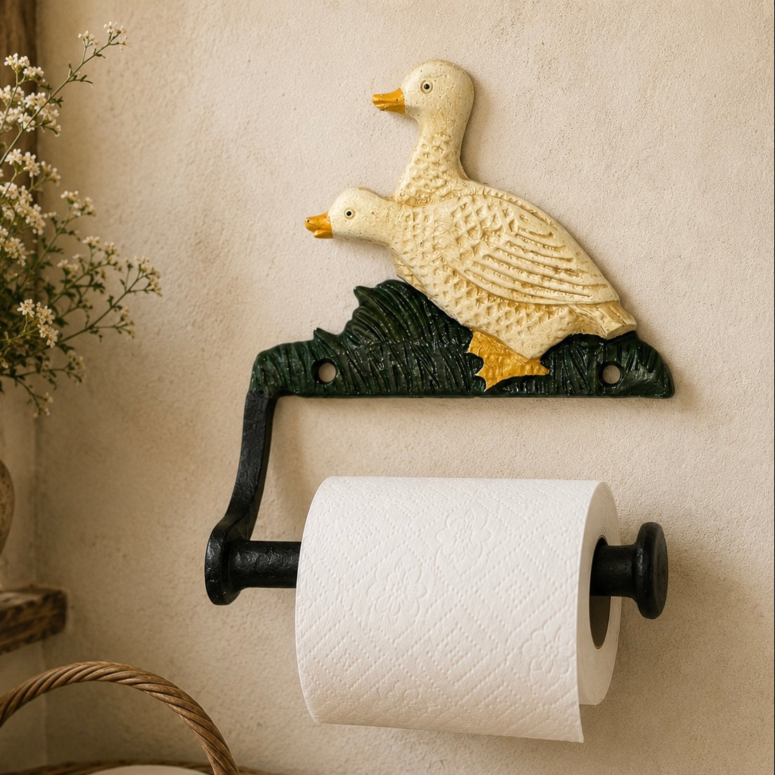 Decorative toilet paper holder with duck design on a wall next to a basket of toilet paper and a vase with flowers.
