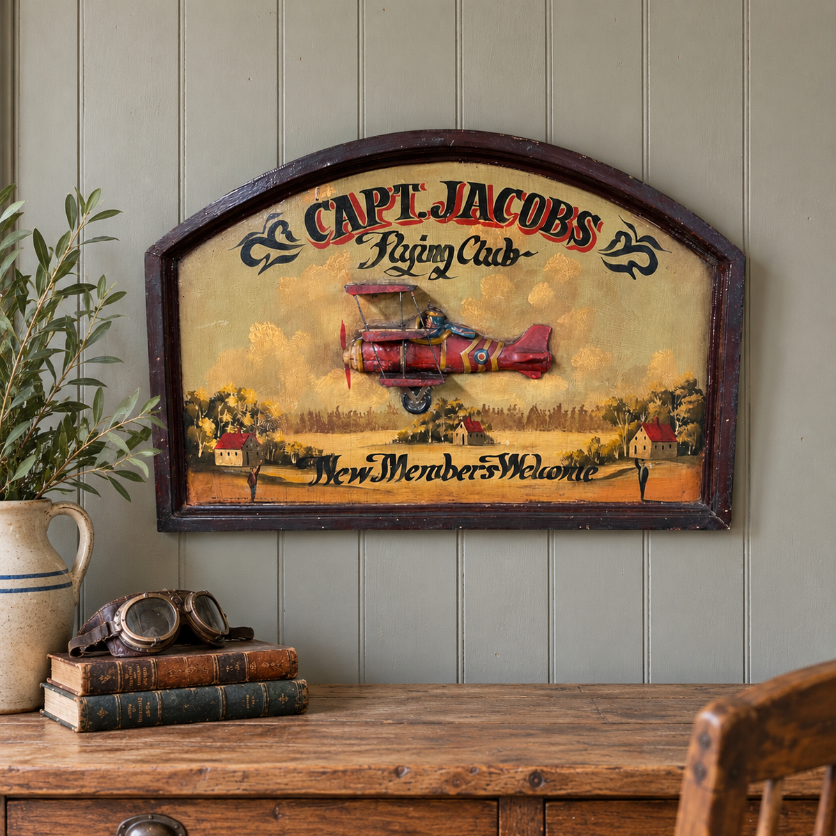 A vintage wooden sign for 'Capt. Jacobs Flying Club' featuring a red biplane and the text 'New Member Welcome,' displayed on a panelled wall above a rustic wooden table with books, a vase, and aviator goggles.