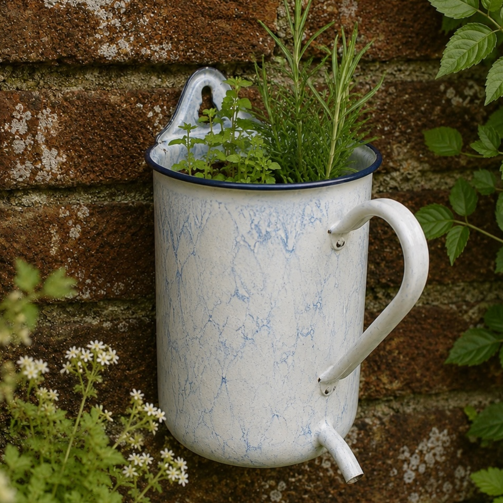 A vintage-style enamel garden planter shaped like a white and blue marbled enamel mug, mounted on a rustic brick wall and filled with green herbs and plants.