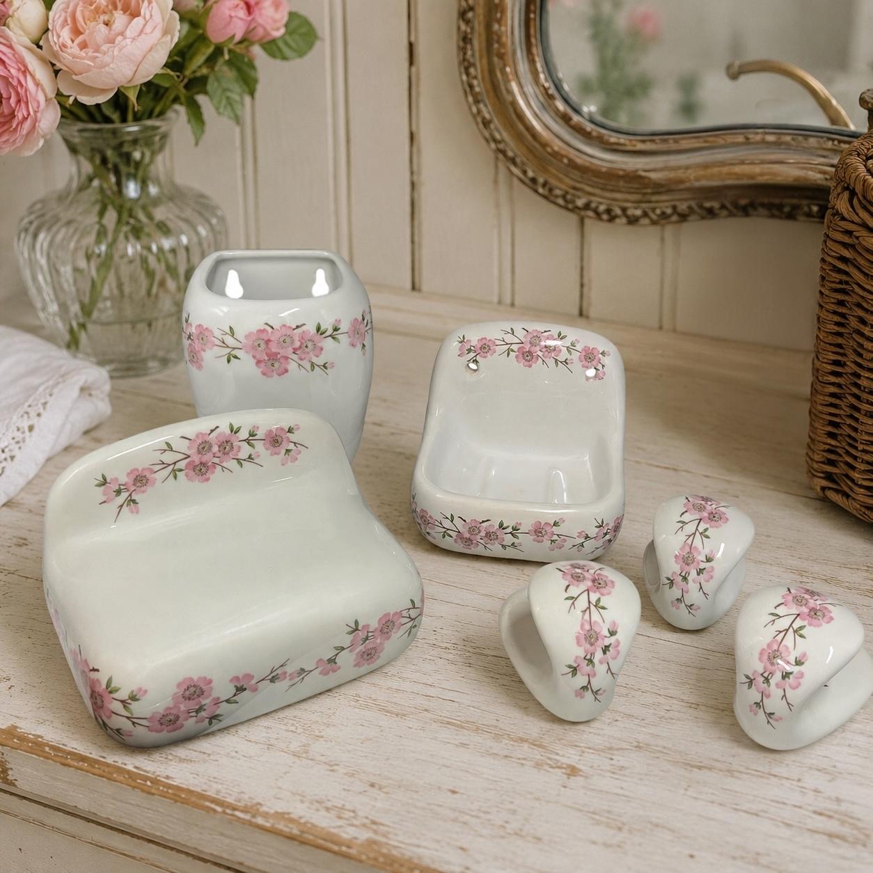 A coordinated set of glossy white ceramic bathroom accessories with pink floral borders, arranged on a distressed white wooden surface. The set includes a rectangular soap dish, a small bowl, two small cup holders, and a round soap dish, styled with a glass vase of pink roses and a mirror in the background.