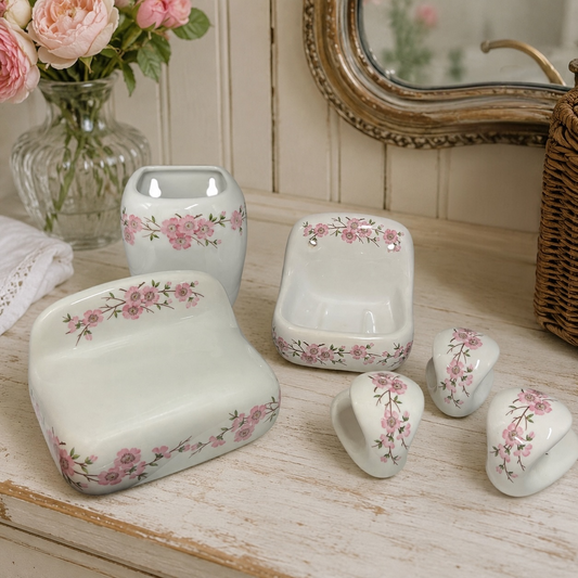 A coordinated set of glossy white ceramic bathroom accessories with pink floral borders, arranged on a distressed white wooden surface. The set includes a rectangular soap dish, a small bowl, two small cup holders, and a round soap dish, styled with a glass vase of pink roses and a mirror in the background.