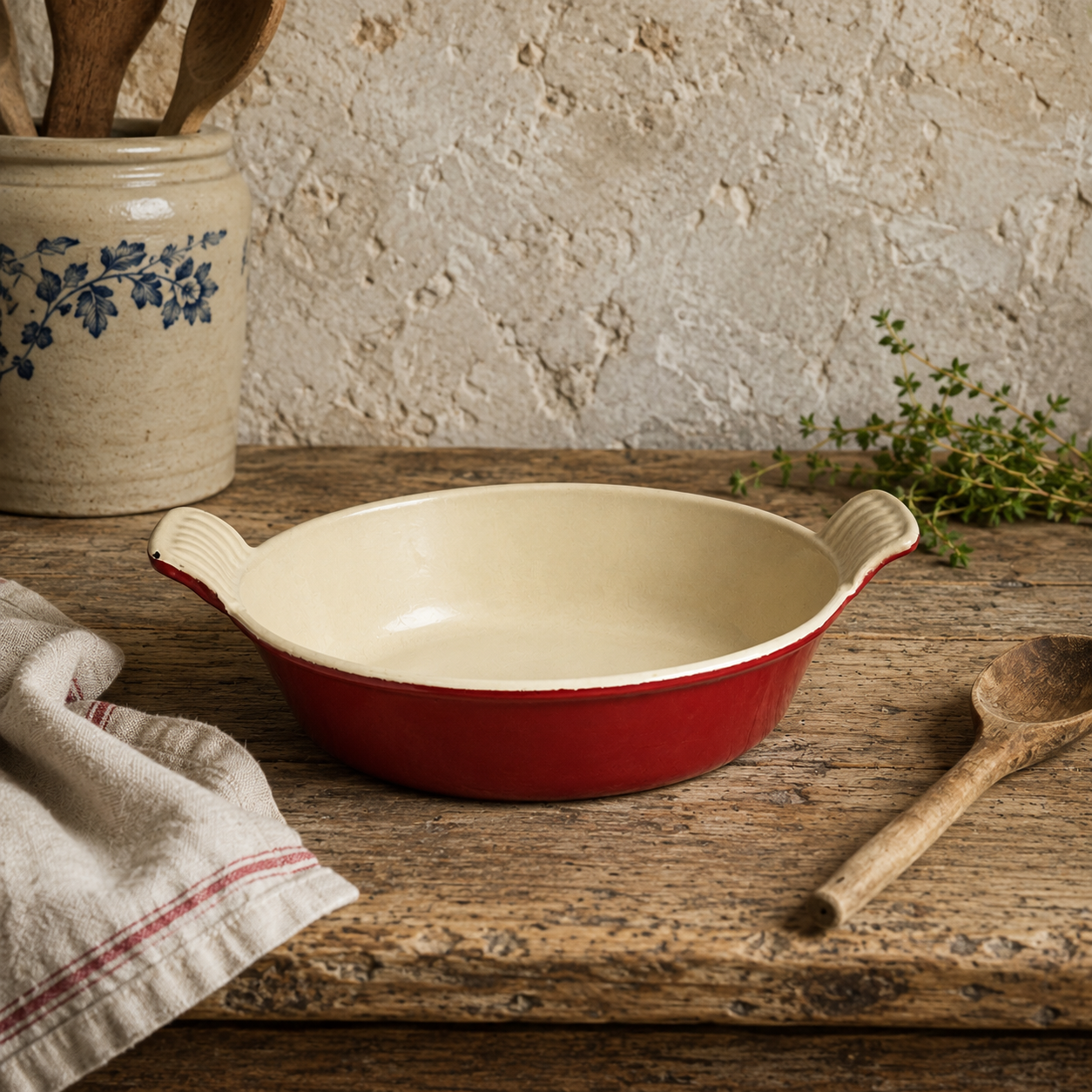 A round red and cream ceramic baking dish with side handles, styled on a rustic wooden table with a wooden spoon, a striped cloth napkin, and a ceramic jar in the background.