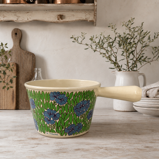 Vintage Le Creuset cast iron fondue pan in rare 1970s floral design, with blue flowers and green leaves, styled on a rustic French kitchen table.