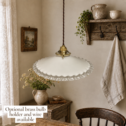 A white glass pendant light with a scalloped edge and a brass top cap hanging in a rustic, cozy room setting. A text overlay in the bottom left corner reads 'Optional brass bulb holder and wire available'.
