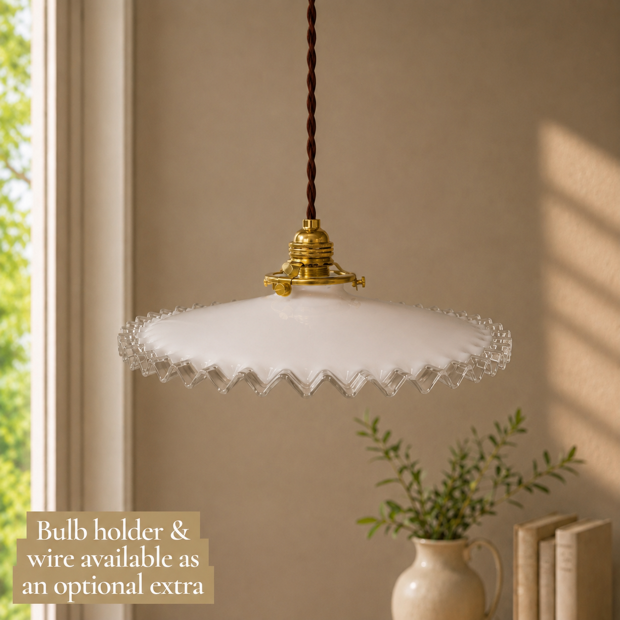 A vintage-style pendant light with a scalloped white glass shade and gold brass fixture, hanging in a warm beige room with a window and a small plant in the background. A text overlay in the bottom left reads 'Bulb holder & wire available as an optional extra'.